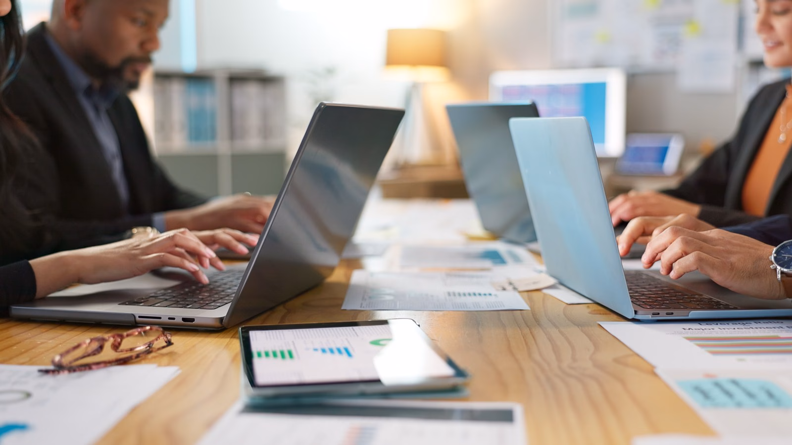 Colleagues use laptops and paper reports around a shared office table.