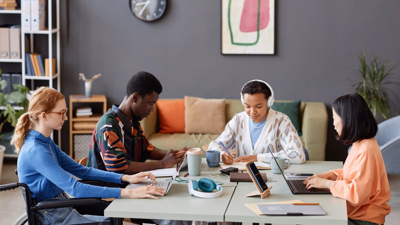 Four colleagues, including a wheelchair user, work together around a table with laptops and notes in a bright office.
