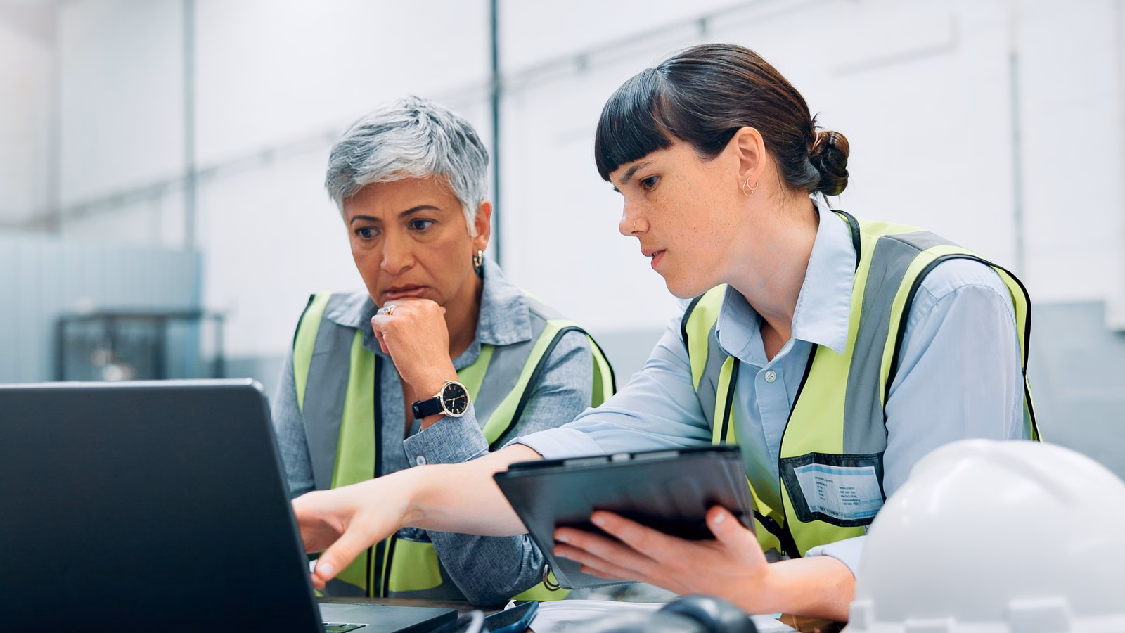 Two manufacturing colleagues in high-visibility clothing review production information on a laptop and tablet.
