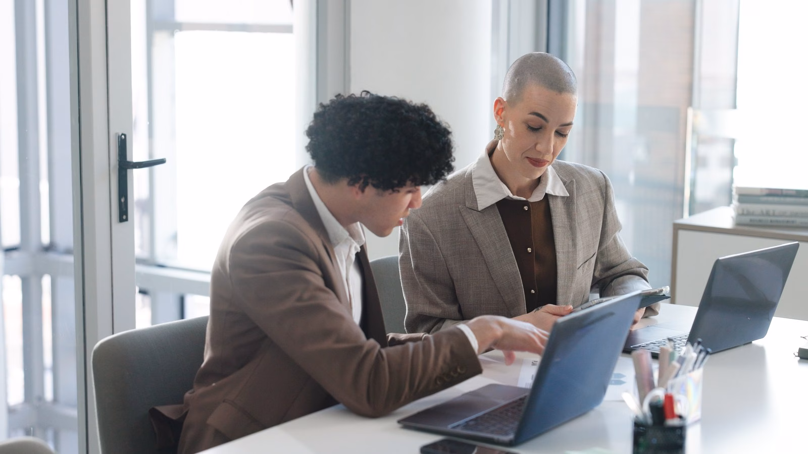 Two colleagues review information together using a tablet and laptops in a bright office.